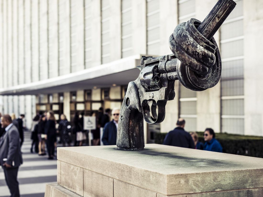 Eine Skulptur eines Revolvers mit verknotetem Lauf steht auf einem Podest vor einem Gebäude, während sich Menschen in der Nähe versammeln.