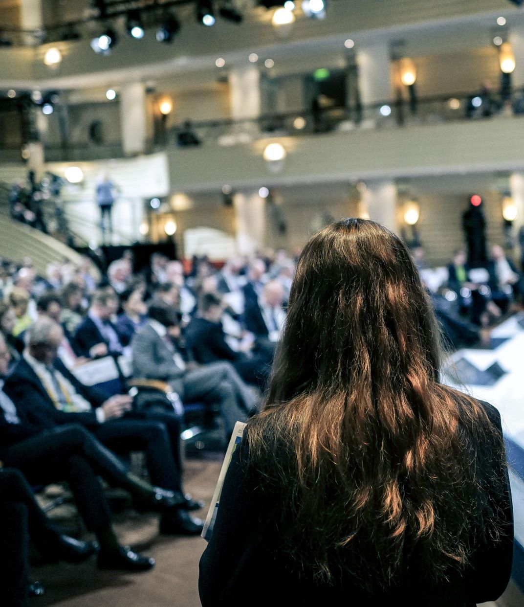 A person with long hair stands next to the main stage with their back to the camera, with the audience of the Munich Security Conference visible in the background.